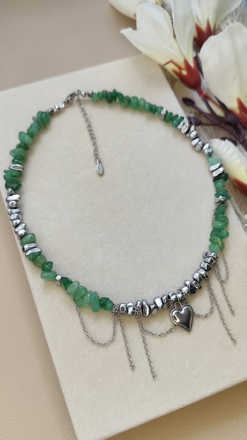 Green beaded bracelet with silver spacers, heart charm, and chain details displayed on a cream card next to white flowers